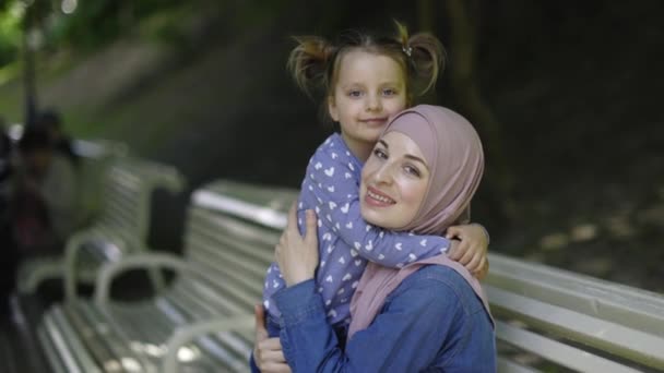 Langzame beweging van gelukkige moslim familie, moeder en dochter, veel plezier in het park. Charmante jonge dame in hoofddoek, zitten in het park op de bank samen met haar schattige kind meisje, knuffelen elkaar. — Stockvideo