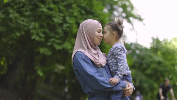 Okouzlující muslimská matka v hidžábu, drží svou roztomilou dcerku v rukou v zeleném parku a užívá si volného času, dotýká se nosy a usmívá se. Zpomalený pohyb maminky a holčičky, procházka v parku. — Stock video