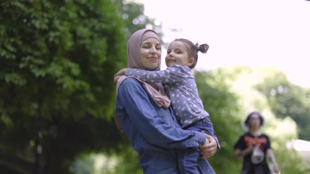 Slow motion van aangename mooie jonge moslim moeder dragen casual jeans kleding en beige hijab, vasthouden aan handen haar schattige dochtertje en knuffelen elkaar tijdens het lopen in het park. — Stockvideo
