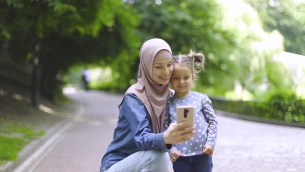 Elég boldog fiatal muszlim anya hidzsáb és aranyos kislánya, hogy szelfi fotó telefon kamera, vagy hogy videohívás barátja, szabadban a nyári parkban a napsütéses napon. — Stock videók