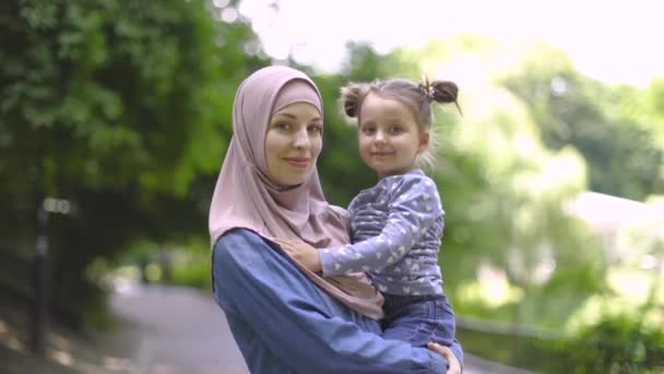 Slow motion van aangename mooie jonge moslim moeder dragen casual jeans kleding en beige hijab, vasthouden aan handen haar schattige dochtertje en knuffelen elkaar tijdens het lopen in het park. — Stockvideo