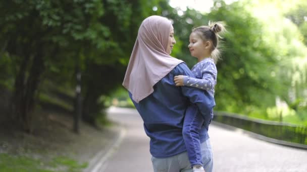 Jonge vrij gelukkige moslim moeder in hijab, spelen met haar kleine schattige dochter in het groene park en houden haar in de armen. Slow motion van vrolijke familie, moeder en kind meisje, wandelen in het park. — Stockvideo