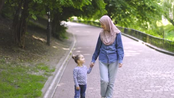 Vooraanzicht van charmante sympathieke jonge moslim moeder en 3-jarige dochter, hand in hand en kijken elkaar, wandelen in het park op prachtige zonnige zomerdag. Gelukkige Arabische familie in het park. — Stockvideo