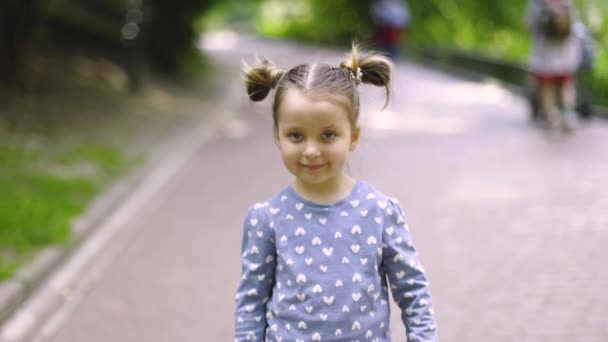 Widok z przodu małej ślicznej dziewczynki, ubranej w niebieską koszulę, chodzącej i pozującej przed kamerą w parku na zewnątrz, machającej ręką i mówiącej cześć lub do widzenia. Spowolnione filmiki 4K. — Wideo stockowe