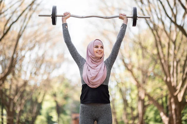 Mujer en ropa deportiva y hijab sosteniendo la barra sobre la cabeza — Foto de Stock