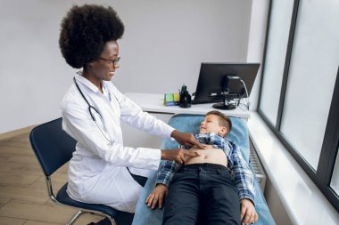 Gülümseyen Afrikalı kadın doktorun portresi elle muayene ediyor, küçük çocuğun karnını inceliyor ve acıtan yerleriyle konuşuyor. Karın ağrısı konsepti.