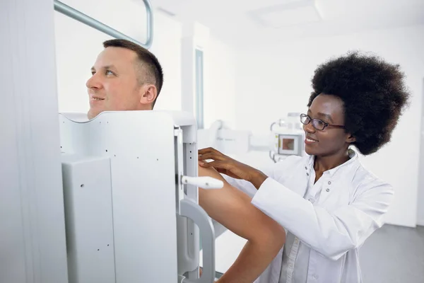 X - ray medical equipment in modern clinic. Female smiling Afro American doctor standing near apparatus, helps male patient during x-ray chest scan. Closeup. — Stock Photo, Image
