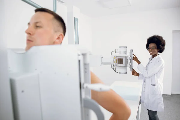 Молодой человек, стоящий в современной больнице во время рентгеновского сканирования грудной клетки, в то время как афроамериканская медицинская техника работает с рентгеновским аппаратом. Сканирование на наличие переломов груди, рака или опухоли. — стоковое фото