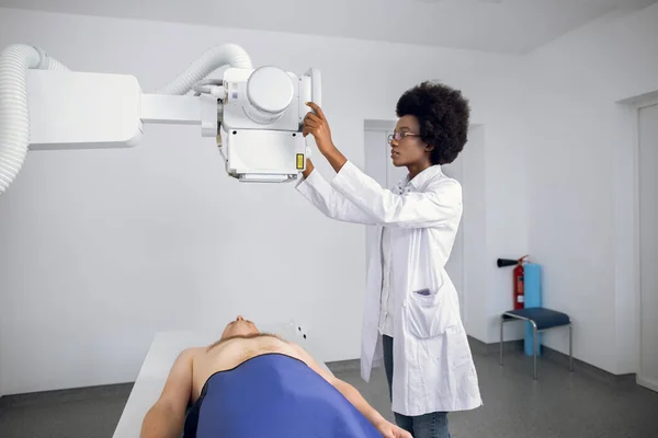 Молодая уверенная афроамериканка-рентгенолог делает рентген молодого пациента лежащего на столе машины. Врач, стоящий рядом с пациентом во время рентгеновской процедуры груди. — стоковое фото
