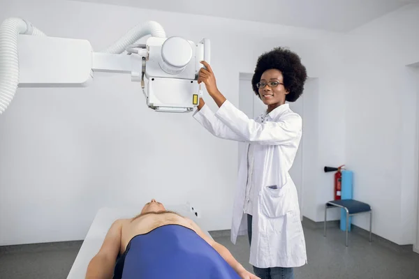 Lächelnde, professionelle afroamerikanische Ärztin in Uniform posiert vor der Kamera, während sie den Bruststrahl eines männlichen kaukasischen Patienten nimmt. Röntgenbild von Lungen und Skelett in moderner Klinik. — Stockfoto