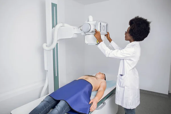 Jovem afro-americana médico radiologista ou técnico, montando máquina para levar pacientes de raio-x. Paciente do sexo masculino deitado durante o procedimento de digitalização. — Fotografia de Stock
