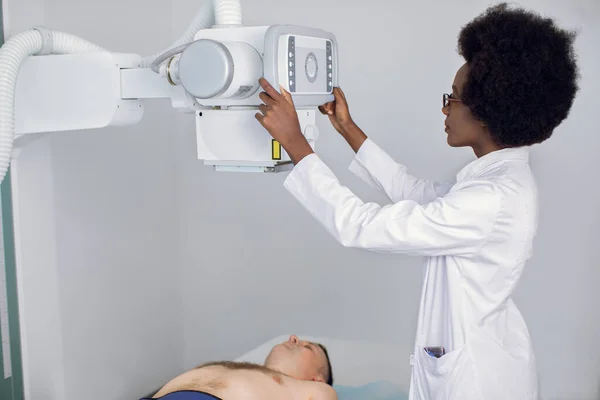 Young african american woman doctor radiologist or technician, setting up machine to take patients x-ray. Male patient lying during scanning procedure. — Stock Photo, Image