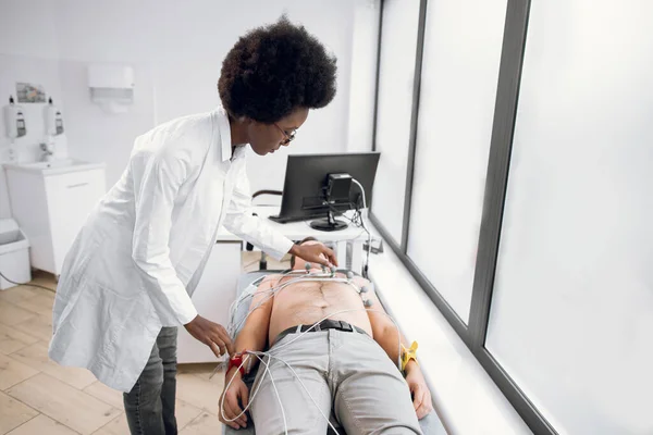 การทดสอบ ECG ของชายหนุ่มที่คลินิกสมัยใหม่ แพทย์หญิงสาวชาวแอฟริกันแพทย์ผู้เชี่ยวชาญด้านหัวใจใส่อุปกรณ์พิเศษขั้วไฟฟ้าเซ็นเซอร์สูญญากาศบนหน้าอกของผู้ป่วยชายผิวขาวก่อนที่จะทํา ecg . — ภาพถ่ายสต็อก