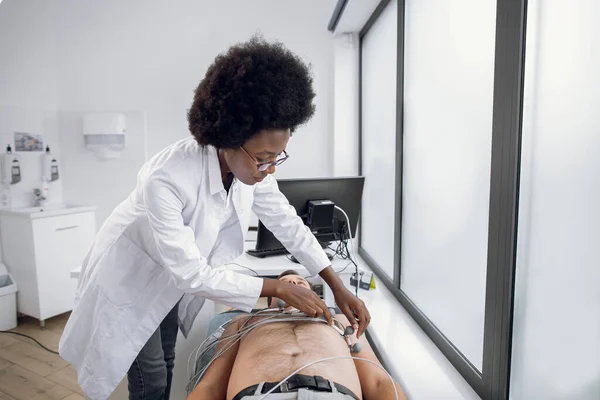 Medicina, saúde e conceito de check-up cardíaco. Jovem afro-americana médica com equipamento de eletrocardiograma fazendo teste de cardiograma para paciente do sexo masculino no hospital, fixando eletrodos no peito do paciente. — Fotografia de Stock