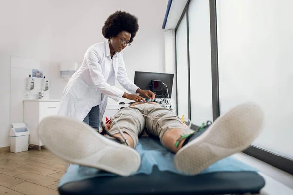 Eletrocardiograma cardíaco para paciente jovem. Jovem médico afro-americano anexa sensores de vácuo no paciente masculino para gravar seu eletrocardiograma na clínica de luz moderna. — Fotografia de Stock