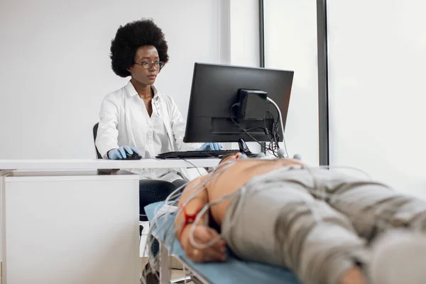 ดร.หญิงสาวชาวแอฟริกันอเมริกันที่มุ่งเน้น นั่งที่โต๊ะและทํางานบนคอมพิวเตอร์ ในขณะที่ทําการทดสอบ ECG ทางหัวใจ สําหรับผู้ป่วยชายในห้องฉุกเฉินของโรงพยาบาล . — ภาพถ่ายสต็อก