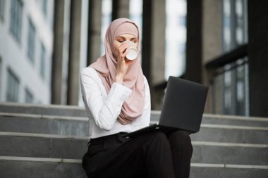 Türbanlı kadın kahve içiyor ve laptopla dışarıda çalışıyor.