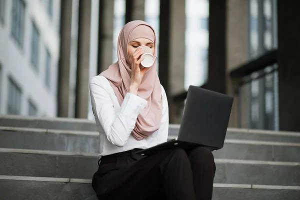 Дама в хіджабі п'є каву і працює на ноутбуці на відкритому повітрі — стокове фото