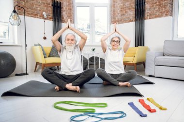 Nilüfer pozisyonunda oturan yaşlı çift elleri başının üstünde.