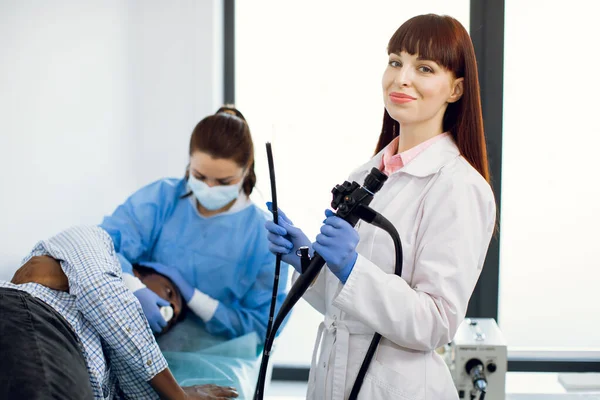 Mujer joven caucásica que sostiene el endoscopio antes de la endoscopia, mientras la enfermera prepara al paciente —  Fotos de Stock