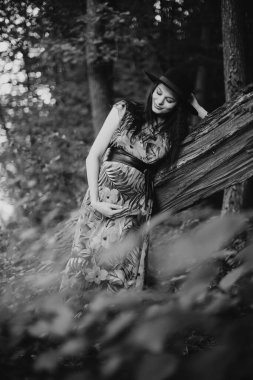 Mutlu ve güzel hamile bir kadının portresi. Göbeğine bakıyor, ağaca yaslanıyor.