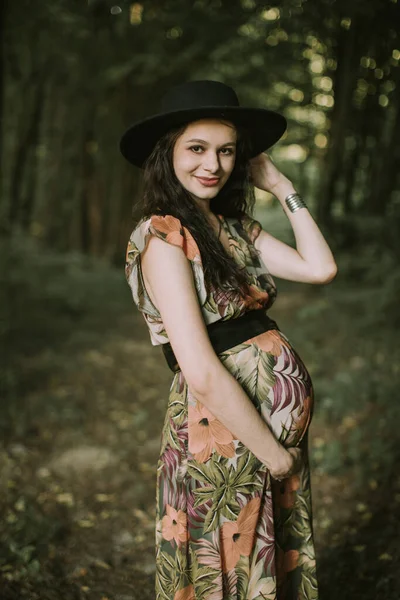 Joven mujer embarazada feliz en vestido elegante y sombrero negro, caminando en el bosque misterioso. —  Fotos de Stock