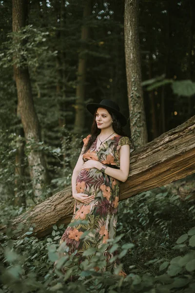 Mujer embarazada en vestido colorido y sombrero negro, de pie cerca del gran árbol caído en el bosque —  Fotos de Stock