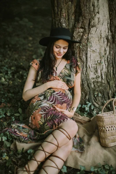 Hermosa mujer embarazada caminando al aire libre en el bosque, sentado debajo del árbol y tocando su vientre. —  Fotos de Stock