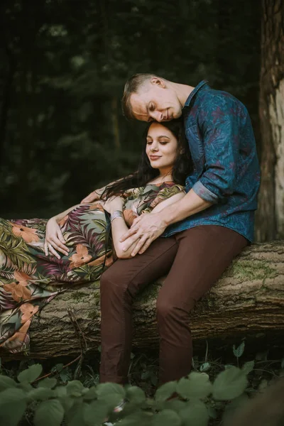 Jeune couple enceinte romantique heureux assis sur l'arbre tombé dans la forêt, étreignant, toucher le ventre — Photo