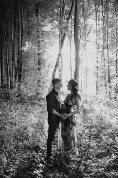 Gelukkig zwangere vrouw en haar man, staande knuffelen elkaar, poseren in het bos tussen de bomen. — Stockfoto
