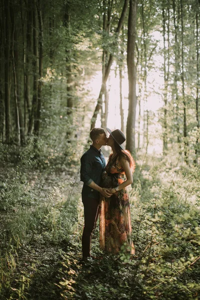 Счастливая беременная женщина и ее мужчина, целующиеся и обнимающиеся, гуляющие по лесу — стоковое фото