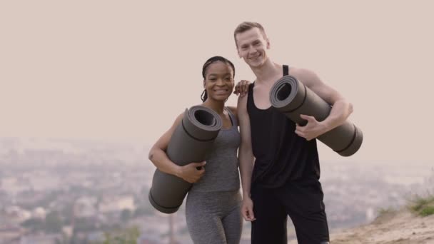 Casal multicultural em roupas esportivas segurando tapetes de ioga, olhando para a câmera e uns aos outros — Vídeo de Stock