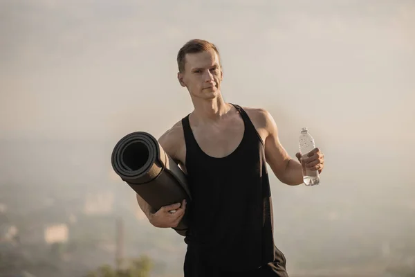 Άντρας αθλητής κρατώντας μπουκάλι νερό και στρώμα γιόγκα σε εξωτερικούς χώρους — Φωτογραφία Αρχείου