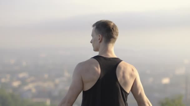 Widok z tyłu muskularny człowiek relaksujący się na wysokim wzgórzu, podnosząc ramiona do nieba — Wideo stockowe