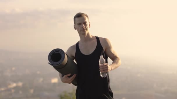 Elinde yoga minderi ve bir şişe su tutan yakışıklı adam kameraya gülümsüyor. — Stok video
