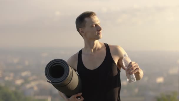 Joven hombre en forma en ropa deportiva sosteniendo estera de yoga y agua potable — Vídeos de Stock