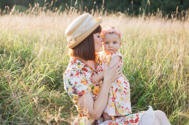 Anne yaz tarlasında çimenlerde oturuyor, kollarında tatlı bir kız çocuğu, nazikçe bebek yanağını öpüyor.