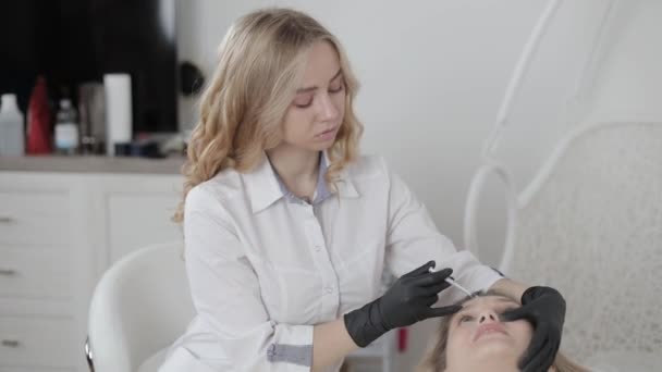 Mãos de cosmetologista feminino fazendo injeção de botox entre as sobrancelhas no rosto de uma mulher bonita. Correção de dobras mímicas no salão. Envelhecimento facial, rejuvenescimento e hidratação. — Vídeo de Stock