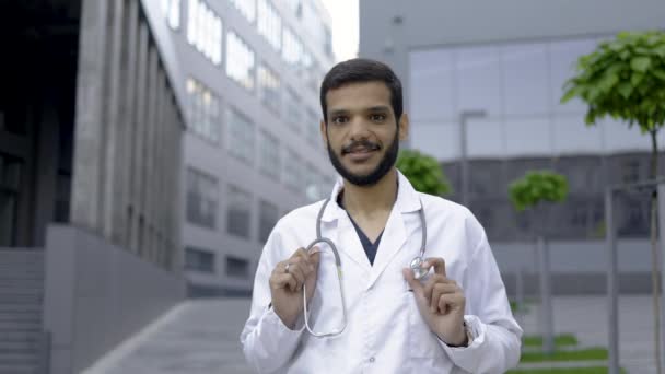 Médecin homme souriant à l'extérieur de l'hôpital, posant en costume médical et manteau blanc, avec stéthoscope — Video