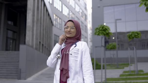 Мусульманская женщина-врач в хиджабе и очках, улыбаясь, позируя перед камерой на улице — стоковое видео