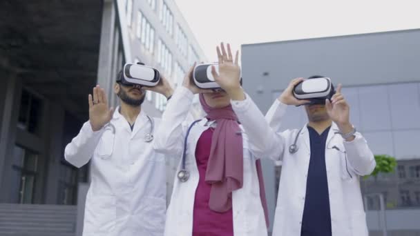 Három fókuszált arab orvos, VR headsetet viselve, mozgó kezekkel a levegőben, kint állva. — Stock videók