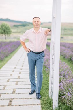 Hafif gömlekli yakışıklı adam lavanta tarlasının arka planında dikilip, yürüyüşün tadını çıkarıyor.