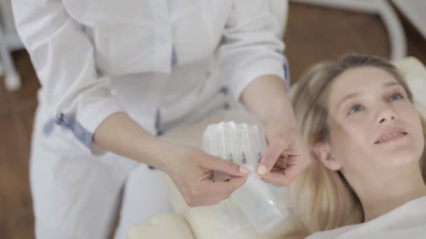 Zugeschnittene Top-Bild des Gesichts der hübschen lächelnden blonden Kaukasierin, bereit, Schönheitsinjektionen, Hautbehandlung zu bekommen, liegend auf dem Sofa in der Kosmetikklinik. Hände der Kosmetikerin halten Satz Spritzen — Stockvideo