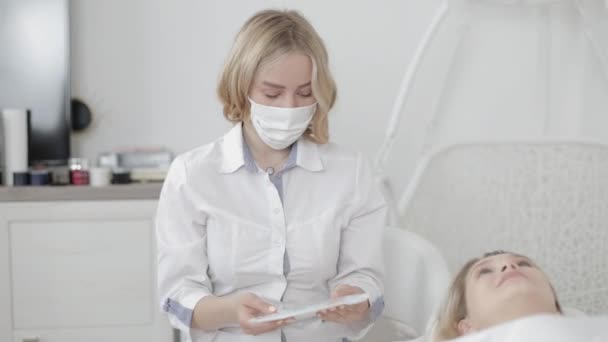 Cirugía estética. Vista del ángulo lateral de la joven mujer caucásica acostada en el sofá y mirando a su médico cosmetólogo, mostrando su conjunto de inyecciones de belleza en la clínica — Vídeo de stock