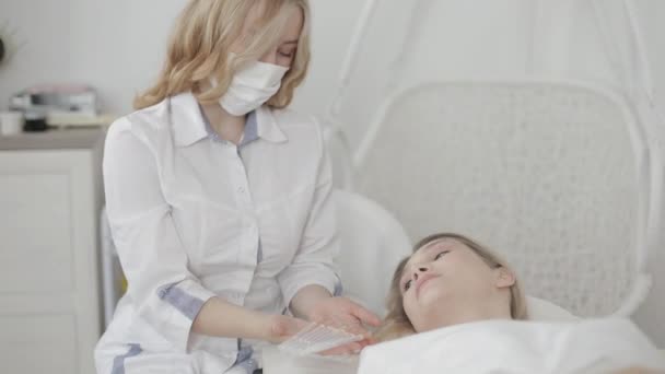 Эстетическая хирургия. Вид сбоку на юную кавказку пациентку лежащую на диване и смотрящую на своего врача-косметолога, показывающую набор инъекций красоты в клинике — стоковое видео