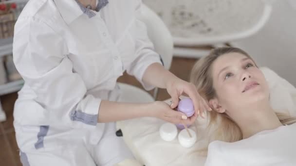 Visiting of beautician. Gorgeous young woman lying on the couch and waiting for relax and beauty procedures in spa center. Hands of female doctor holding bottle of cream. Focus on hands with cream — Stock Video