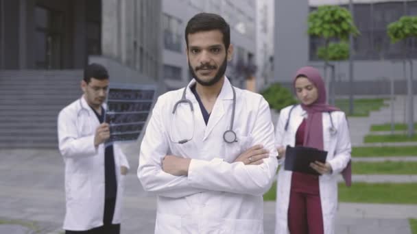 Arabski lub hinduski lekarz przed wielonarodowym zespołem, patrząc w kamerę z skrzyżowanymi ramionami — Wideo stockowe