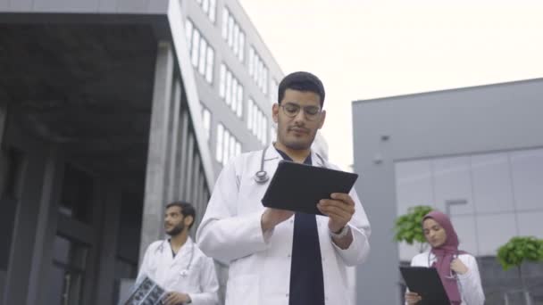 Три молодых врача, работающих с планшетным ПК, рентгеном и пациентами медицинской карты за пределами больницы — стоковое видео
