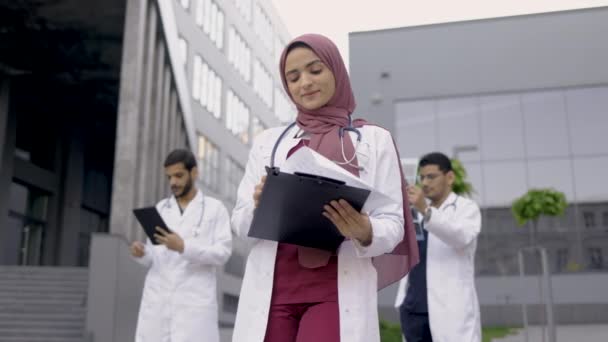 Dokter wanita dalam jilbab menulis catatan di papan klip, berdiri di luar klinik dengan rekan-rekan di belakang — Stok Video