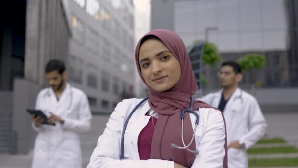 Sonriente médico árabe en hiyab posa al aire libre frente a una clínica moderna con colegas detrás — Vídeo de stock